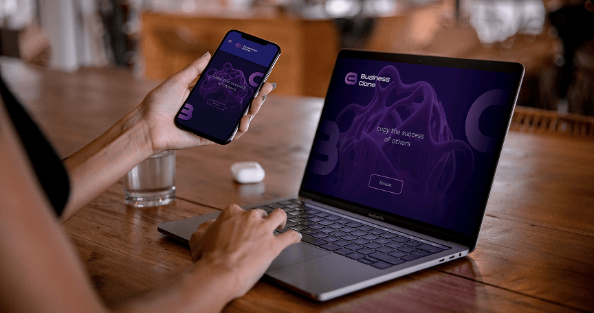 A woman is working on a laptop while holding a smartphone, both displaying the 'Business Clone' app with a purple interface and the text 'copy the success of others.' A glass of water and white wireless earbuds case are on the wooden table beside her.